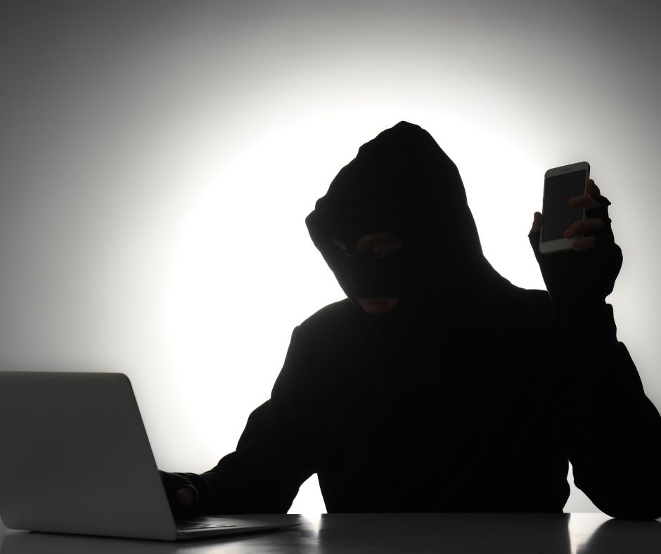 A man in a hoodie holding a phone and working on a laptop